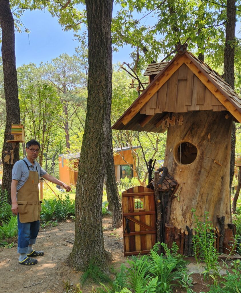 A person in an apron stands beside a wooden structure resembling a small house in a forested area, pointing towards a tree. Lush greenery surrounds the scene.